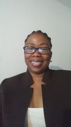 Smiling person in glasses and a dark blazer, standing against a plain wall.