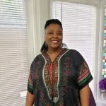 Smiling person in a patterned tunic standing by bright windows with white blinds