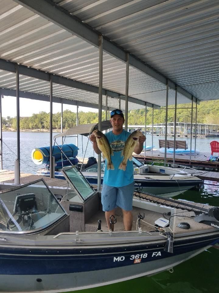 Man holding two large fish on a boat dock, boats in the background. Bright day, blue water.