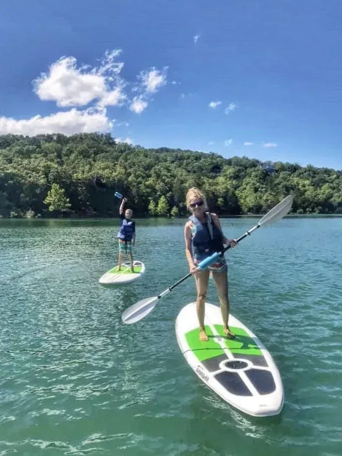 Two people paddleboarding on a lake, surrounded by trees under a blue sky.
