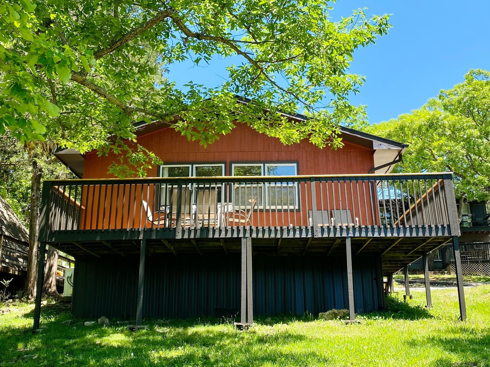 Red cabin with a large deck, surrounded by trees and green grass.