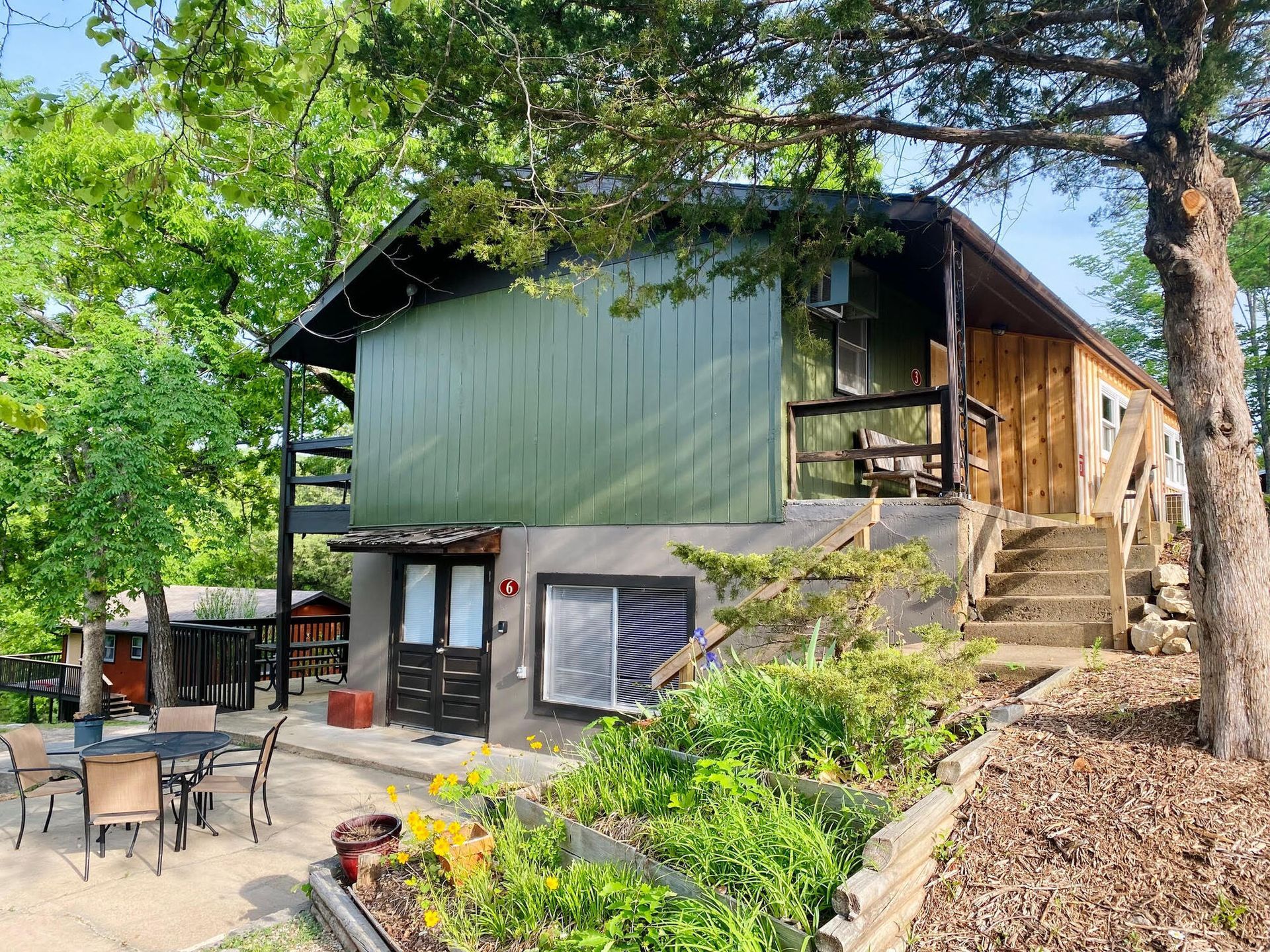 Two-story cabin with green siding, wooden porch, concrete patio with table/chairs, surrounded by trees.