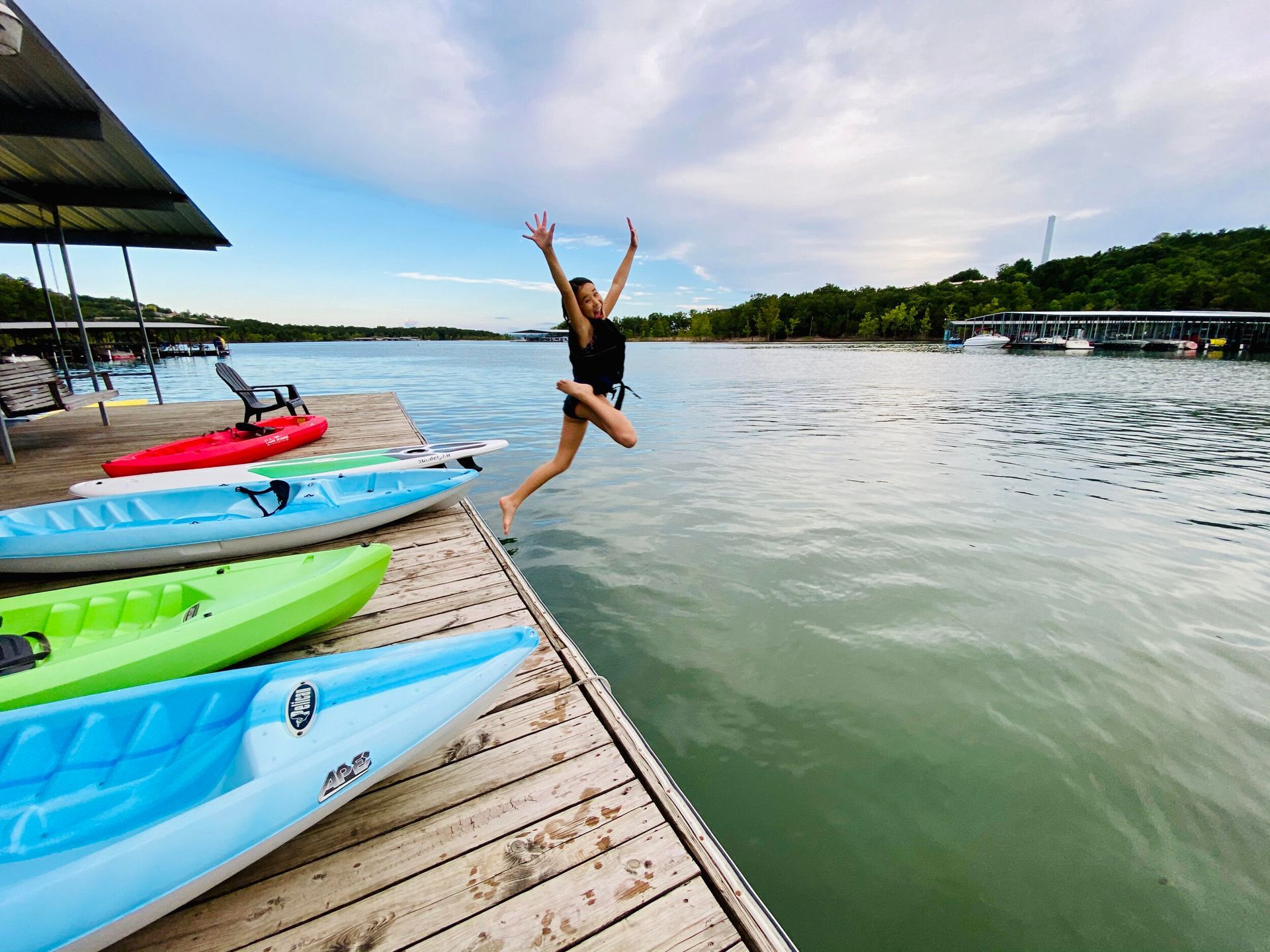 Person jumps from a wooden dock into water near colorful kayaks; sunny day.