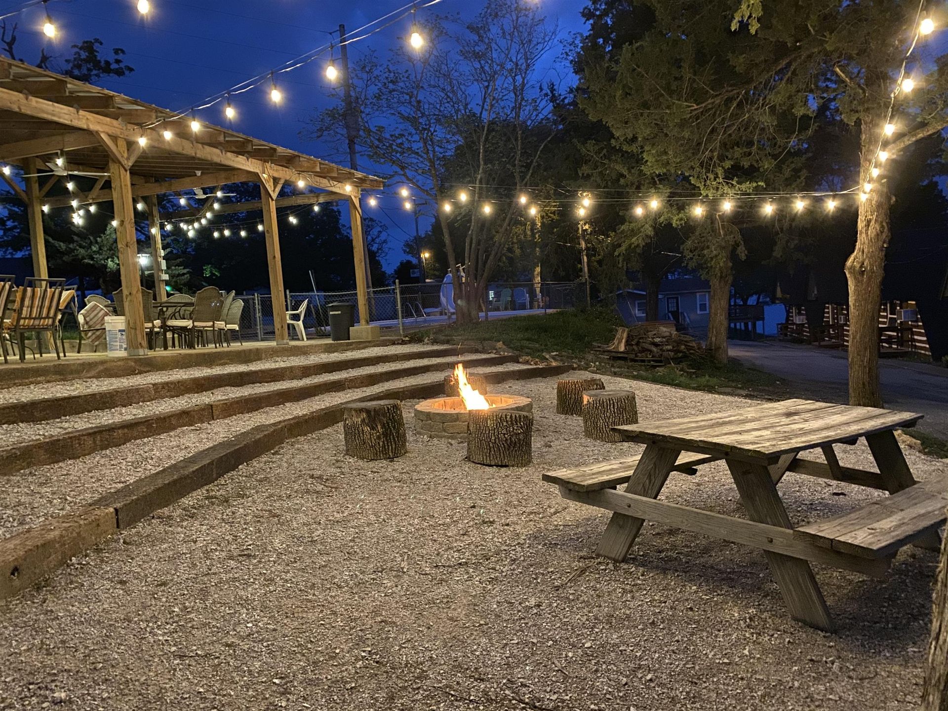 Outdoor event space at night with fire pit, picnic table, and string lights.