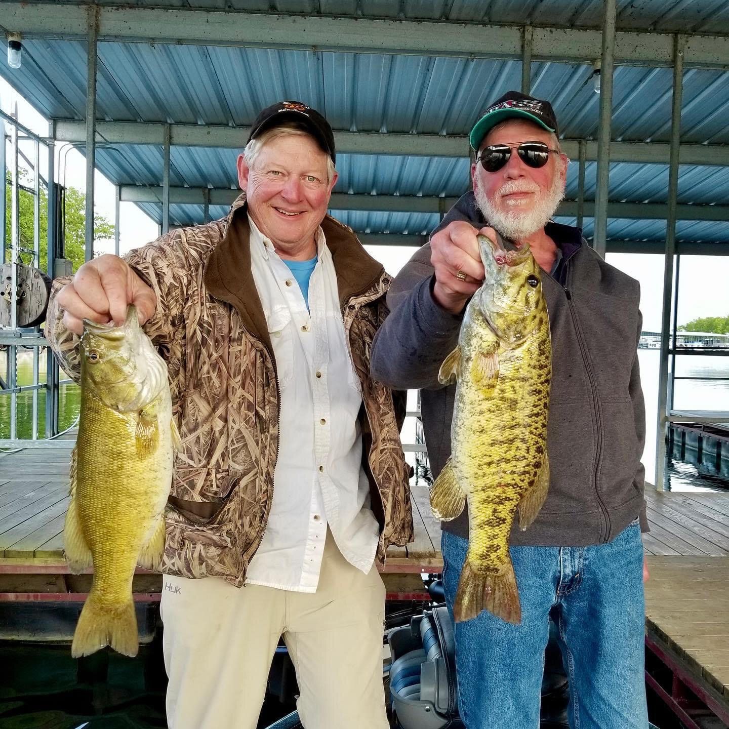 Two men holding up fish they caught, smiling. They are standing near a dock under a metal roof, with water visible.
