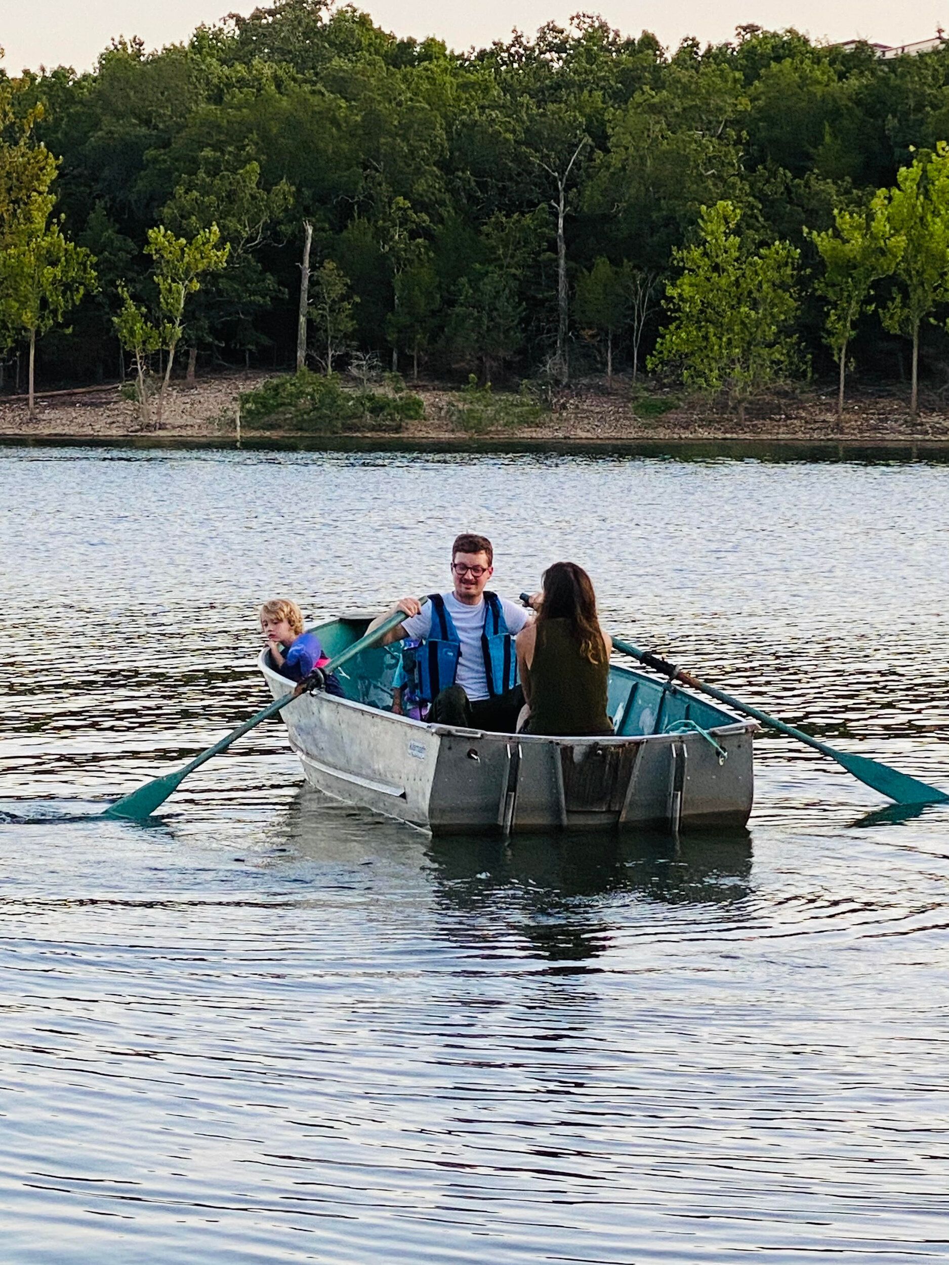 People in a small boat on a lake, surrounded by trees. One person is rowing with blue oars.