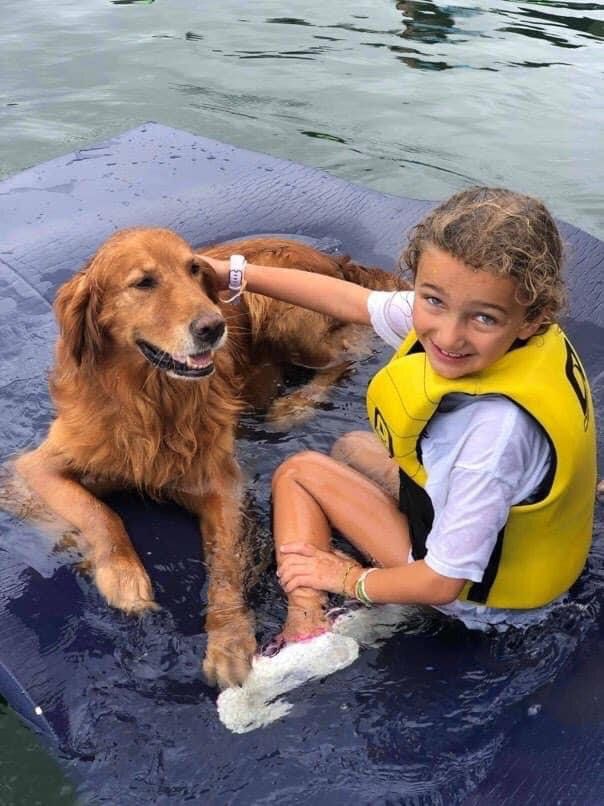 Girl in a life vest and golden retriever dog on a blue floating mat in water, girl petting the dog.