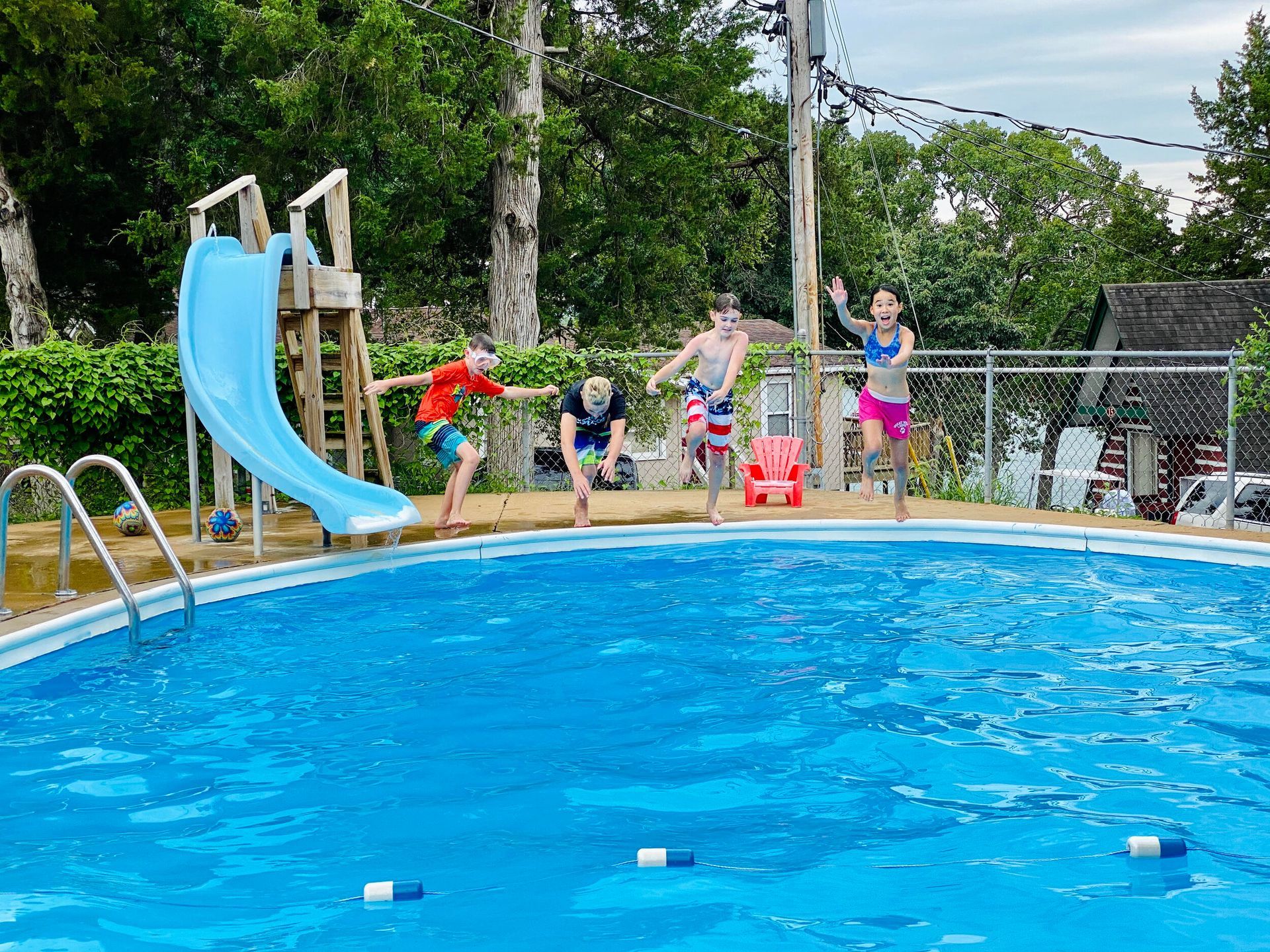 Four people jumping into a blue swimming pool on a sunny day, with a slide visible.