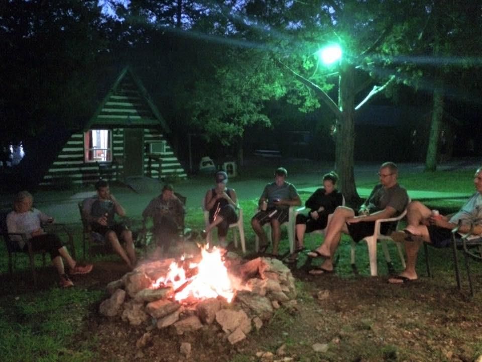 People sit around a campfire at night near a cabin. Firelight illuminates their faces.