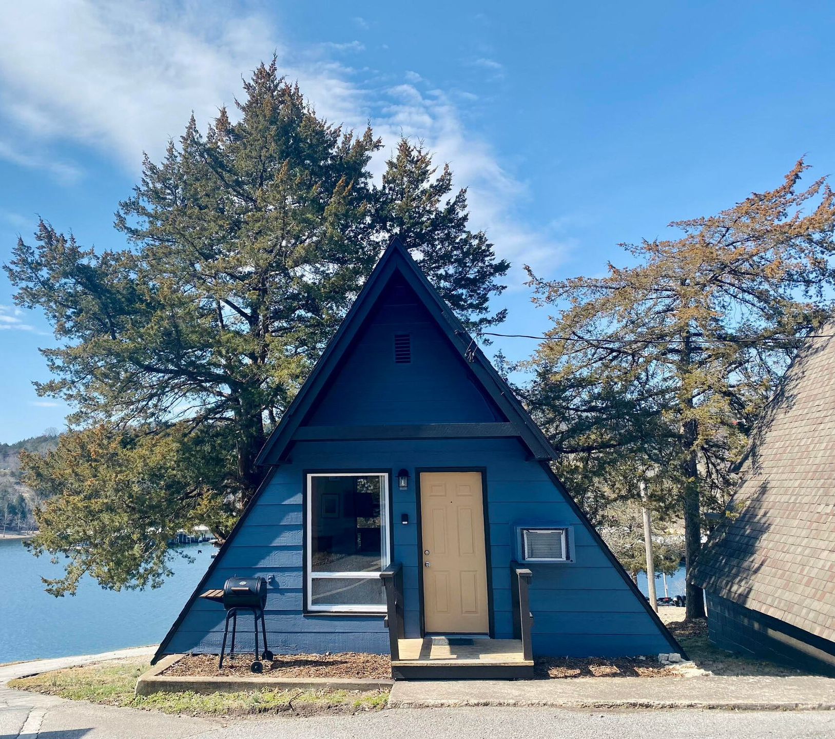 A-frame cabin painted blue, with a door and window, situated near a body of water on a sunny day.