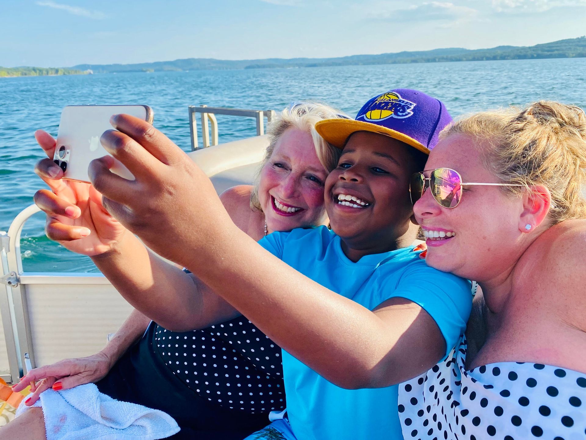 Three people on a boat taking a selfie. Smiling boy wearing a purple cap, women in polka dots. Lake and blue sky.