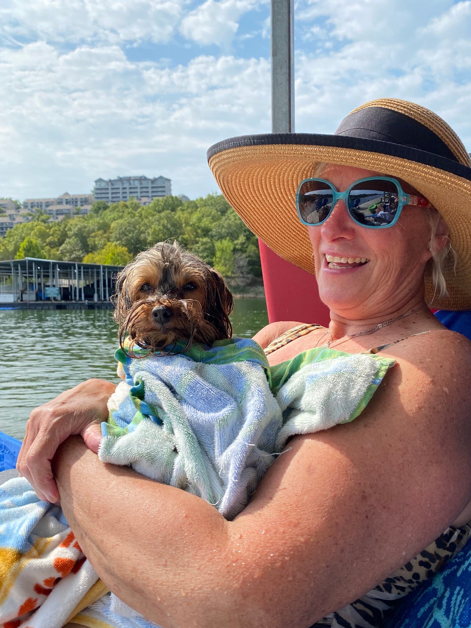 Woman in a straw hat and sunglasses smiles while holding a wet, small dog wrapped in a towel on a lake.