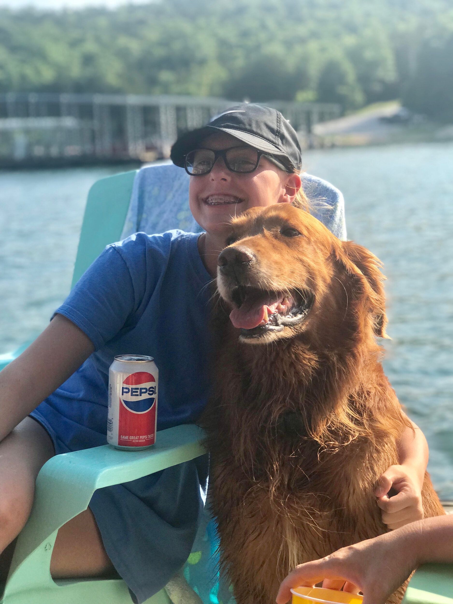 Boy with glasses and golden retriever on a boat, holding a Pepsi. Lake in background.