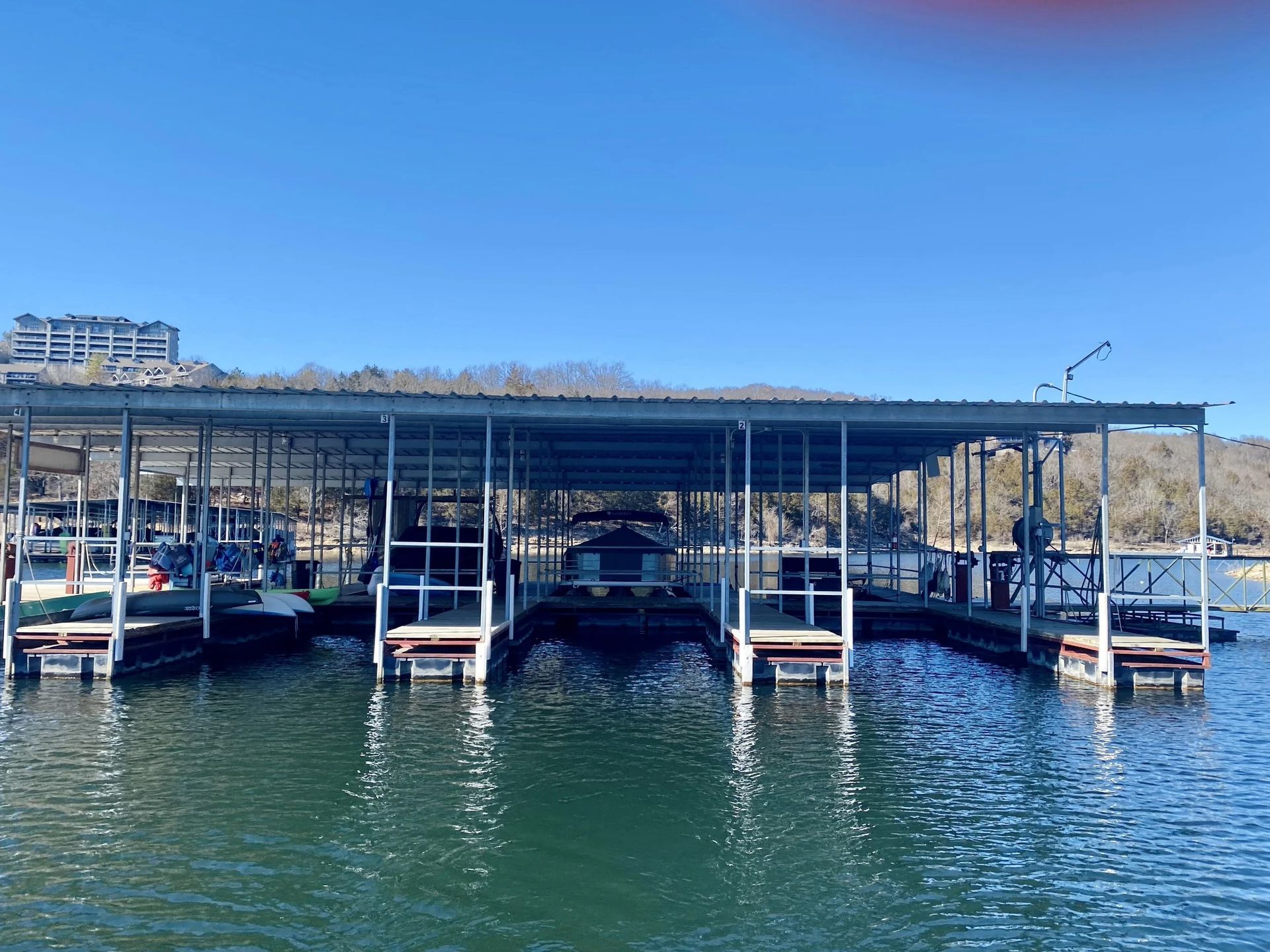 Boat docks on a lake under a metal roof, with boats in slips, blue water and sky.