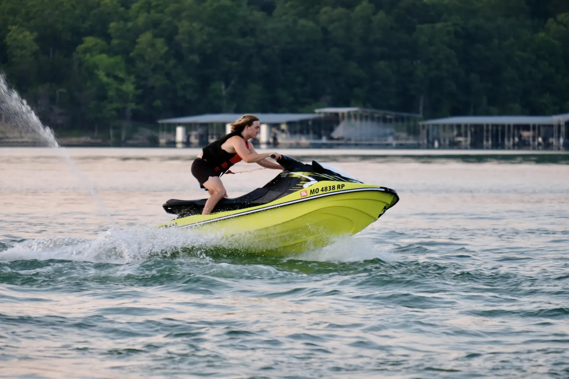 Woman on a yellow jet ski, speeding across water, with a forested shoreline and docks in the background.