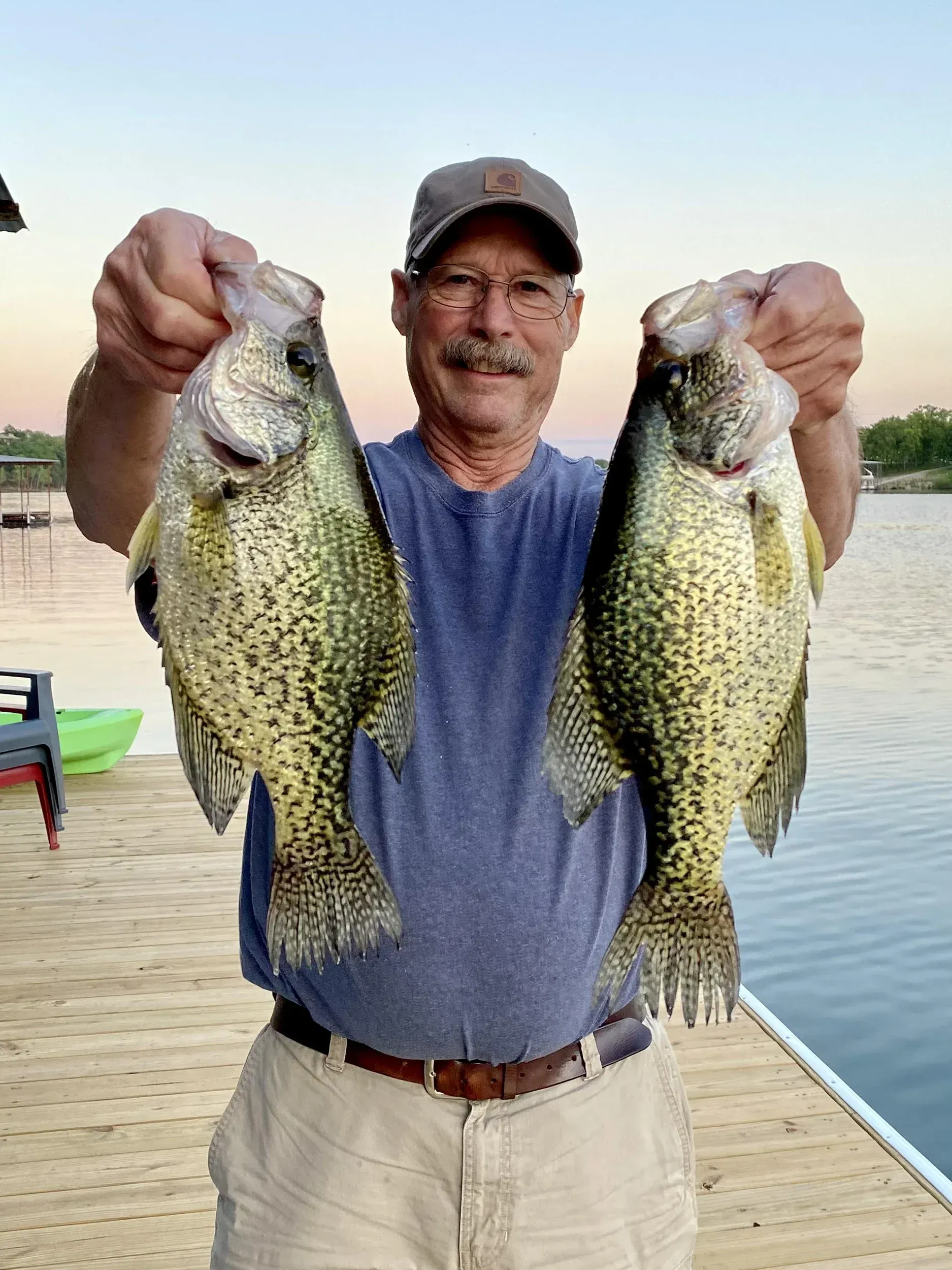 Man holding two spotted fish on a dock near water.