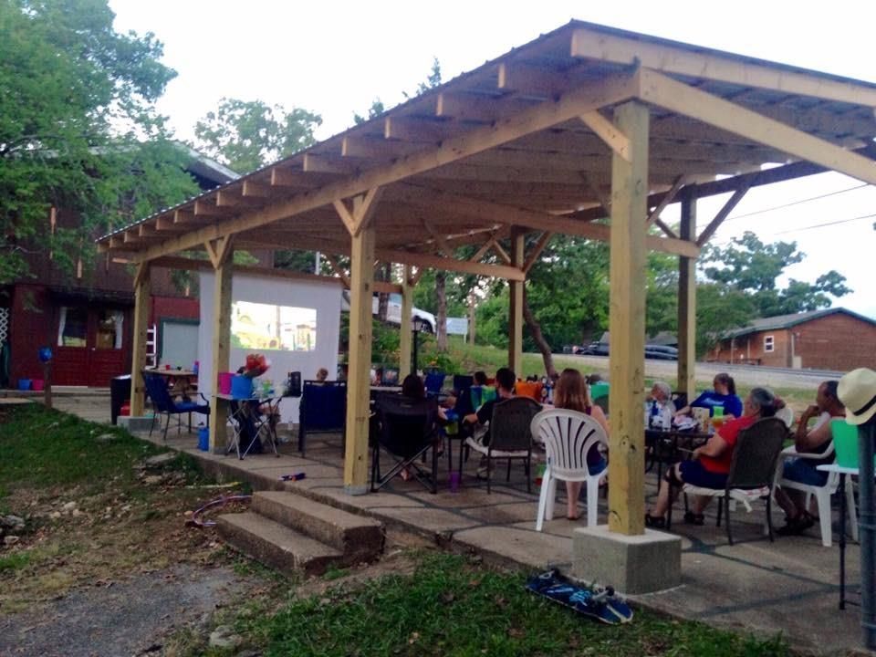 People watching a movie outdoors under a wooden shelter, projected on a screen.