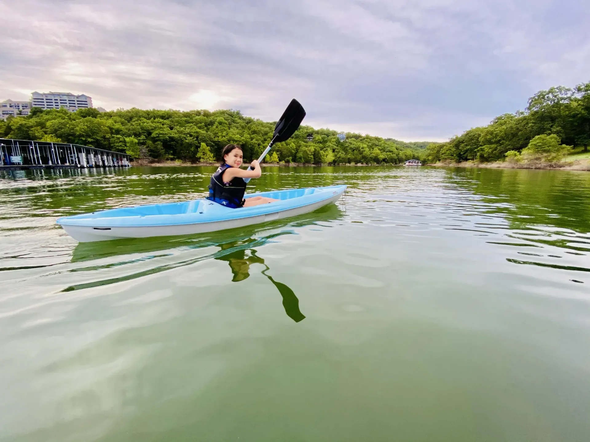 Woman kayaking on a calm lake, paddling with a black oar, surrounded by green trees under a cloudy sky.