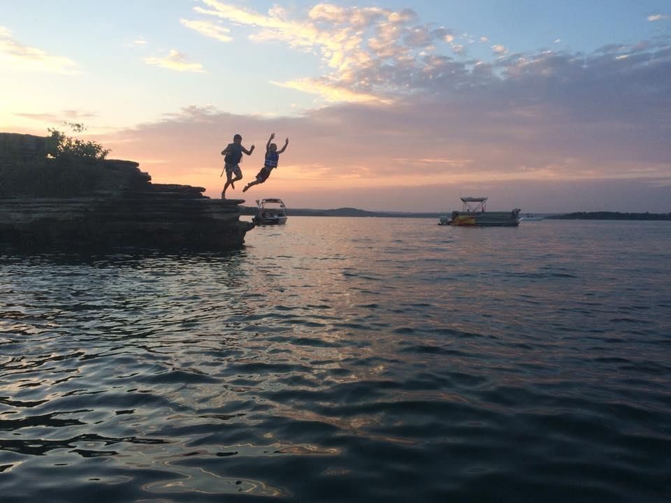 Two people jumping off a rocky cliff into a body of water at sunset.