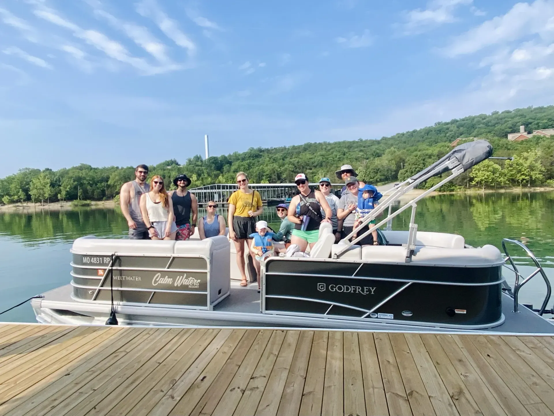 People on a pontoon boat docked on a lake, mountain in the background, blue sky.