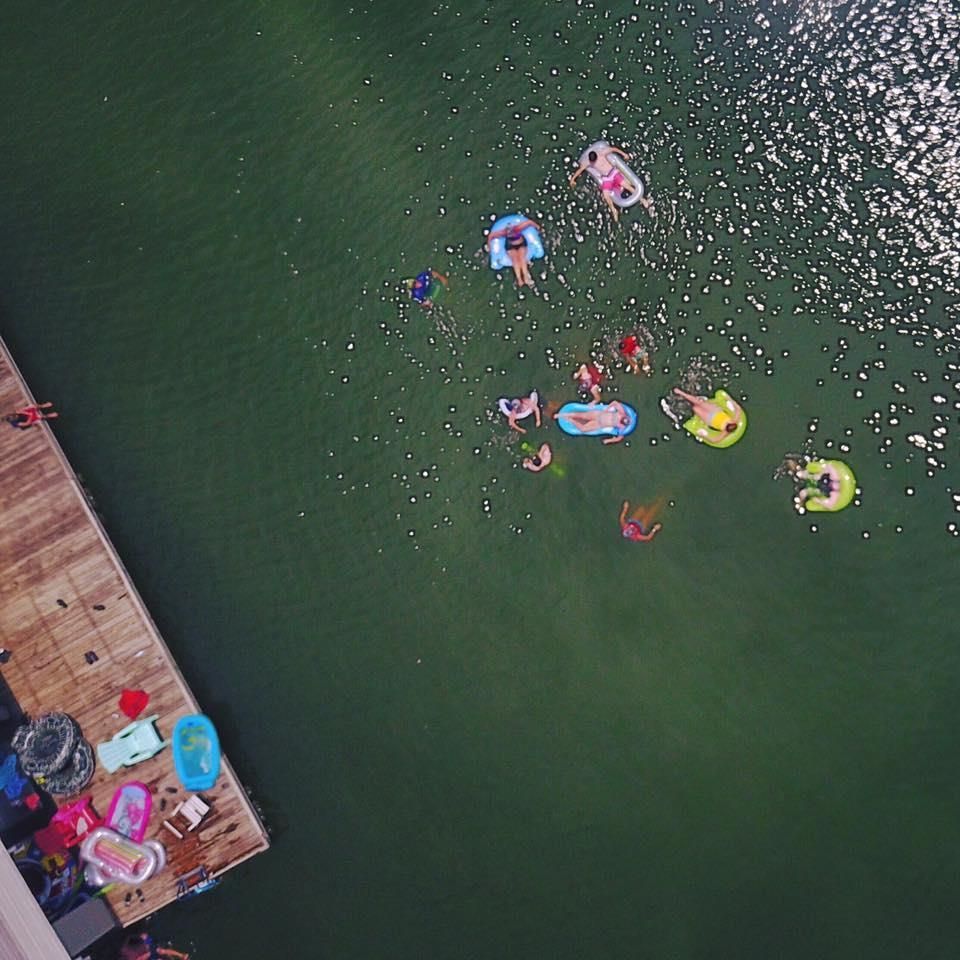Overhead view: People in tubes and swimming near a dock on a green lake; sunlight reflects.