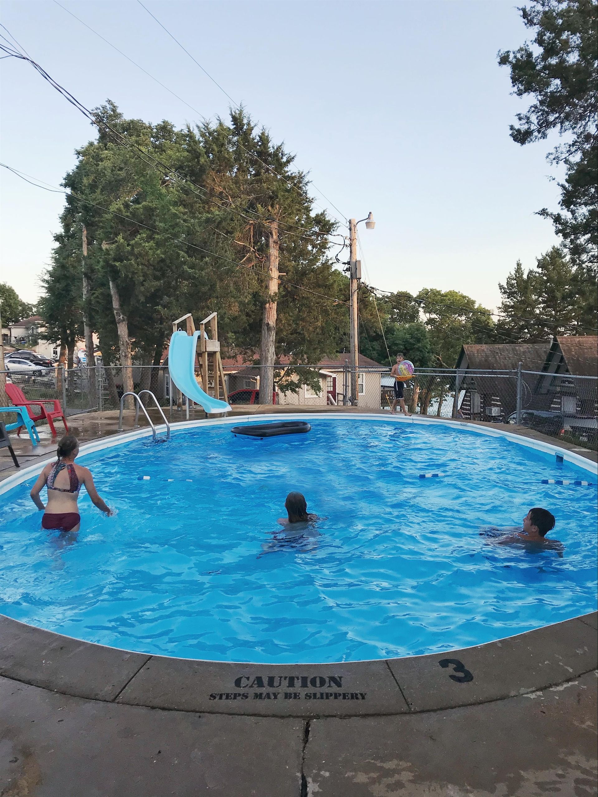 Three people swim in a backyard pool with a slide. Trees and a fence surround the pool.