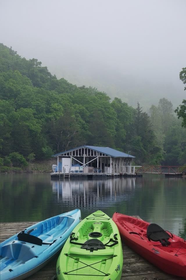 Three kayaks on a dock overlooking a lake with a boathouse and misty, tree-covered hills.