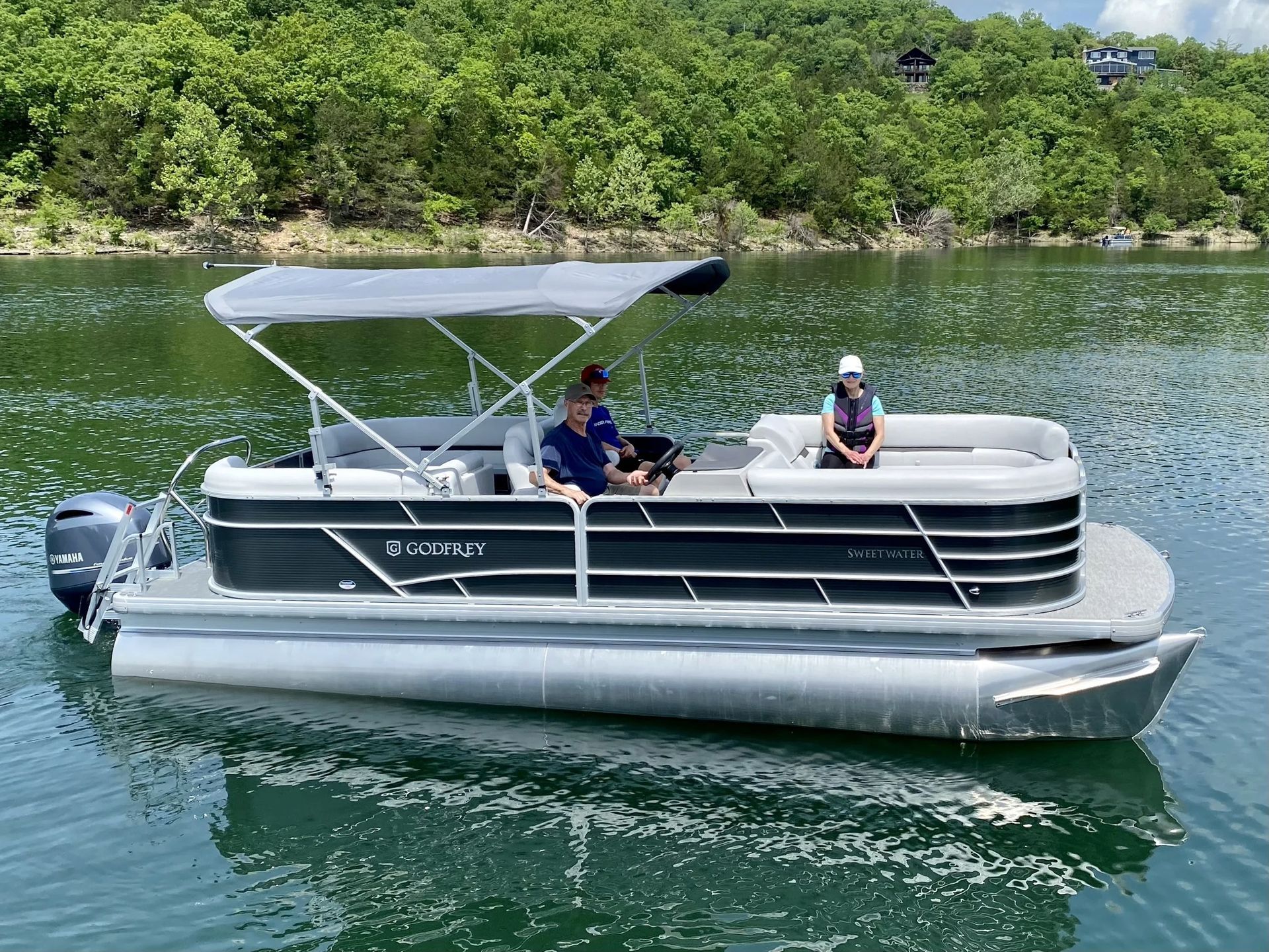 Pontoon boat on lake with three people onboard; sunny day, trees in background.