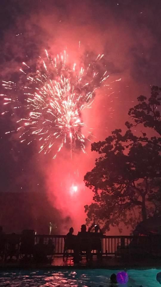 Fireworks burst over a pool at night; silhouettes of people watch from a wooden deck.