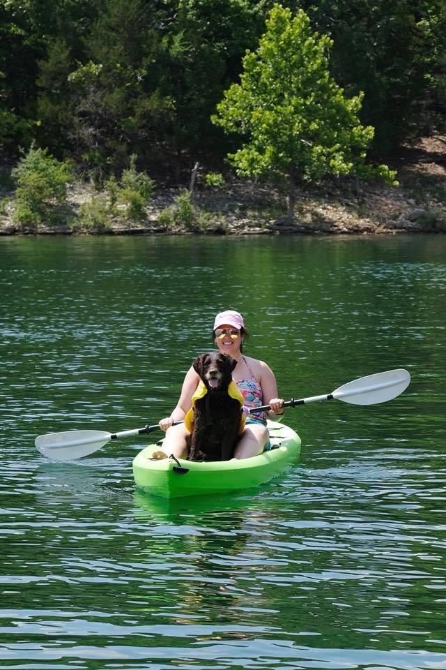 Woman and dog kayaking on a lake. Bright green kayak, blue water, green trees in background, sunny day.