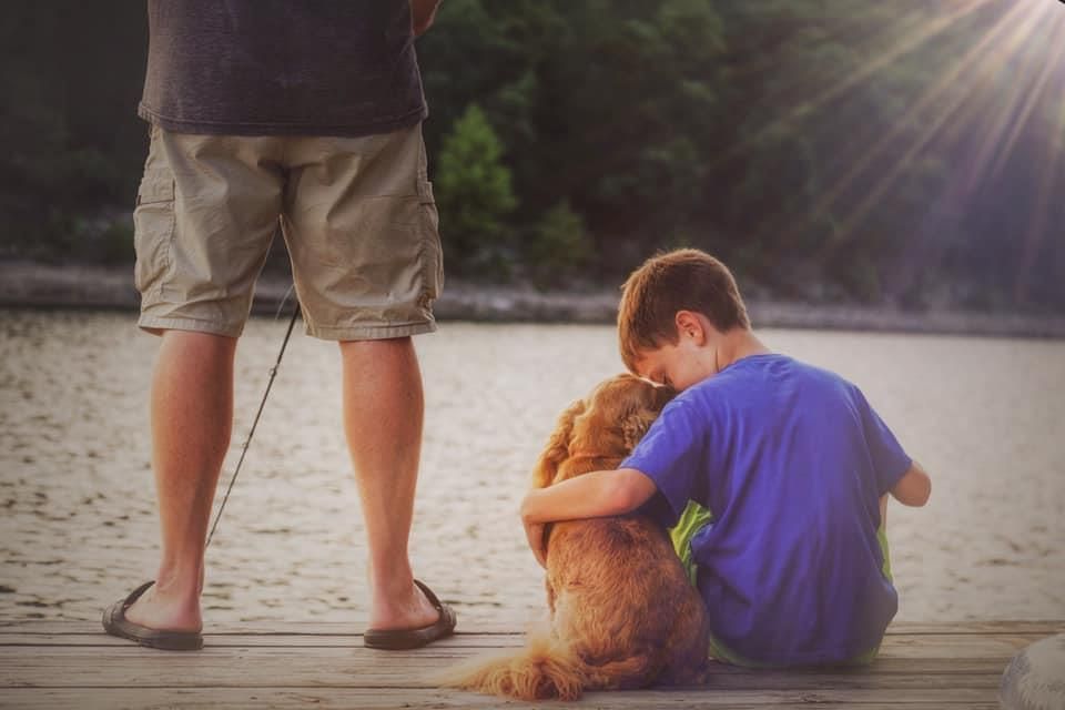Boy hugging dog on a dock, with a person fishing nearby; sunny lakeside setting.