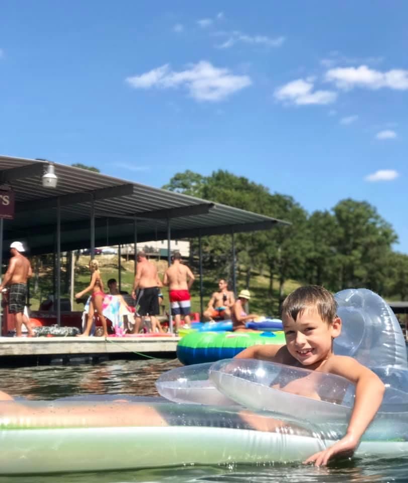 Boy in clear inflatable tube smiles at camera in lake. People relax on dock in background on sunny day.
