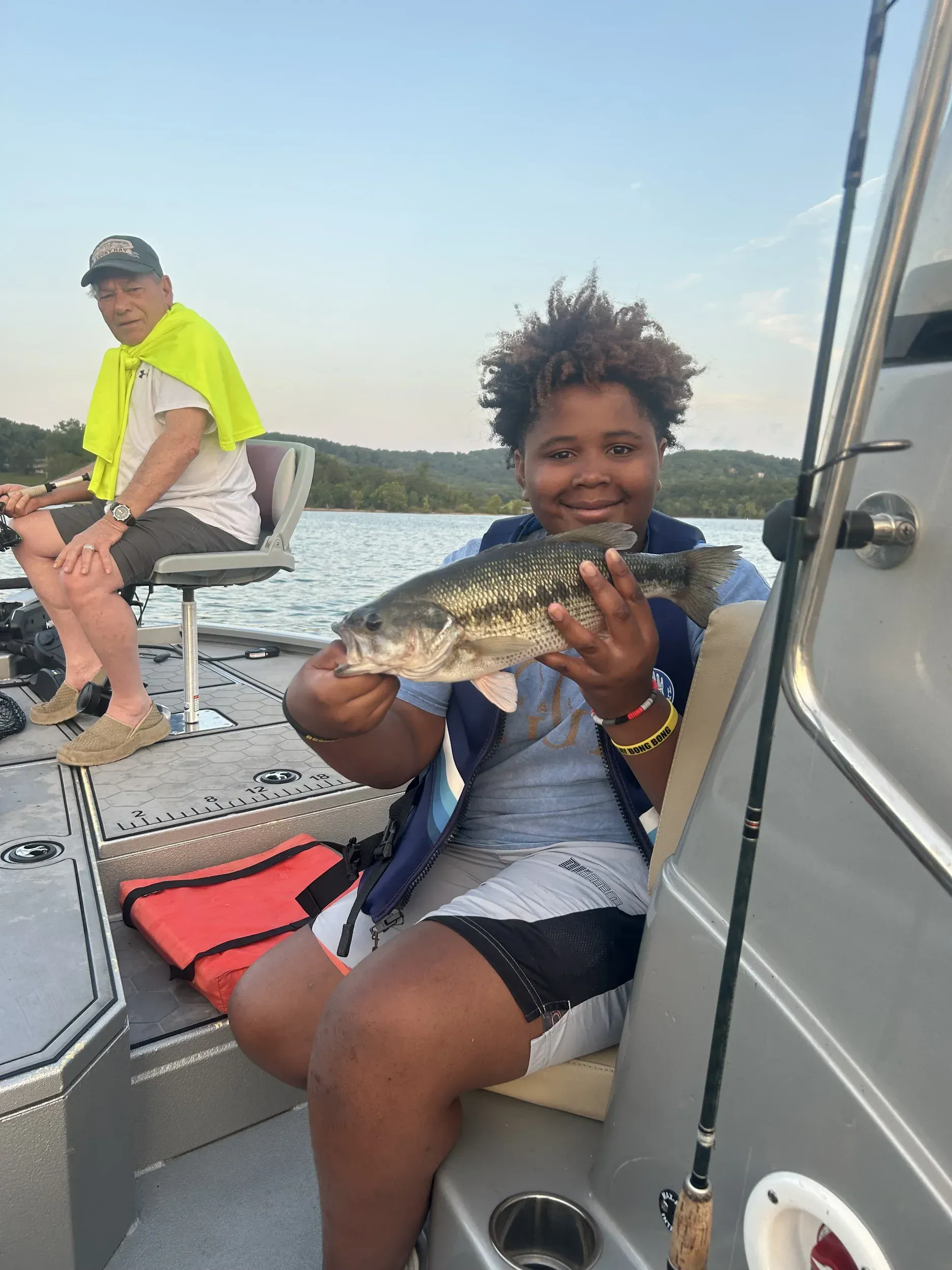 Person on a boat holding a fish, smiling. Another person sits nearby. Lake in background.