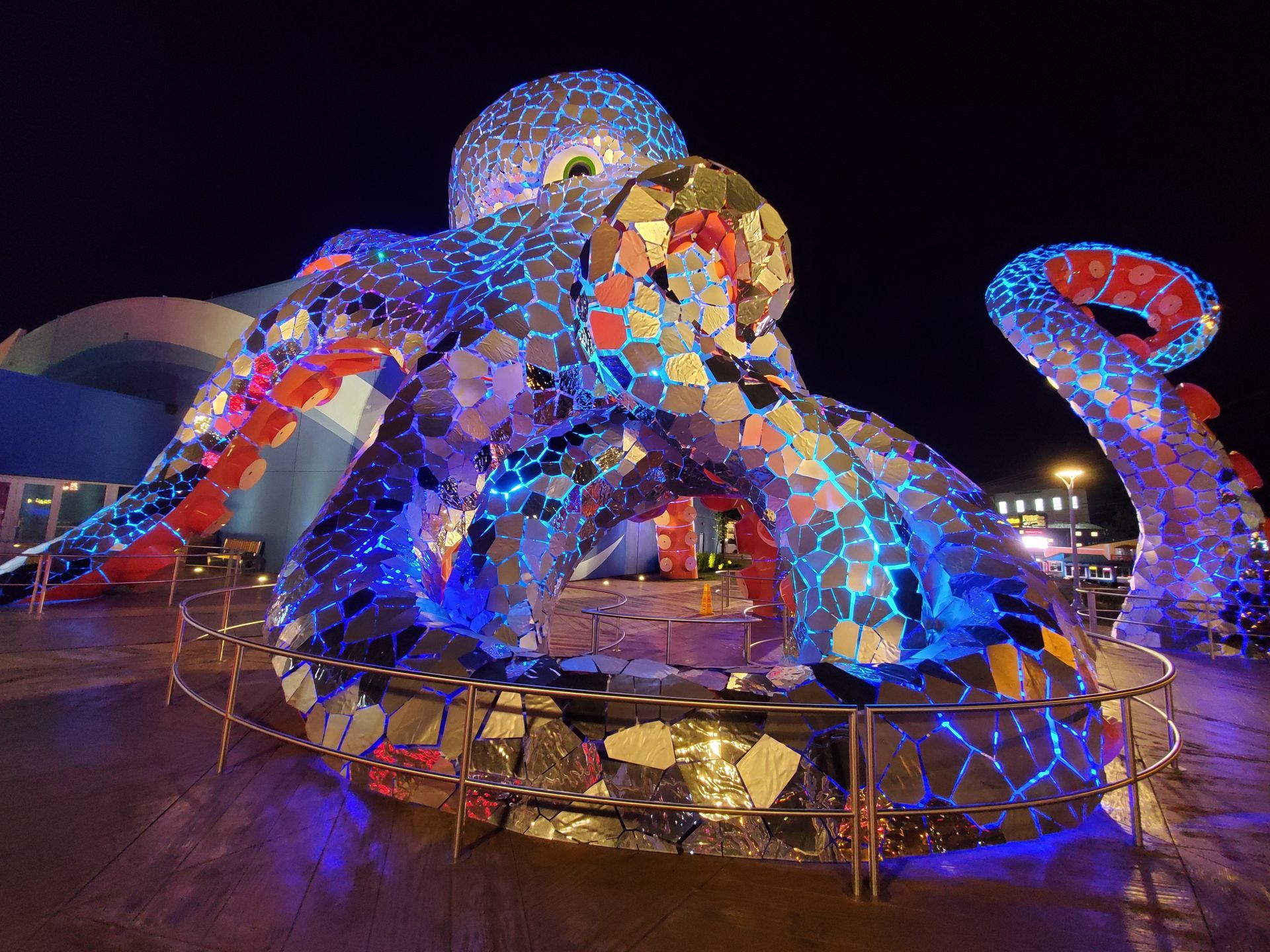 Large, mirrored octopus sculpture illuminated with blue and red lights at night.