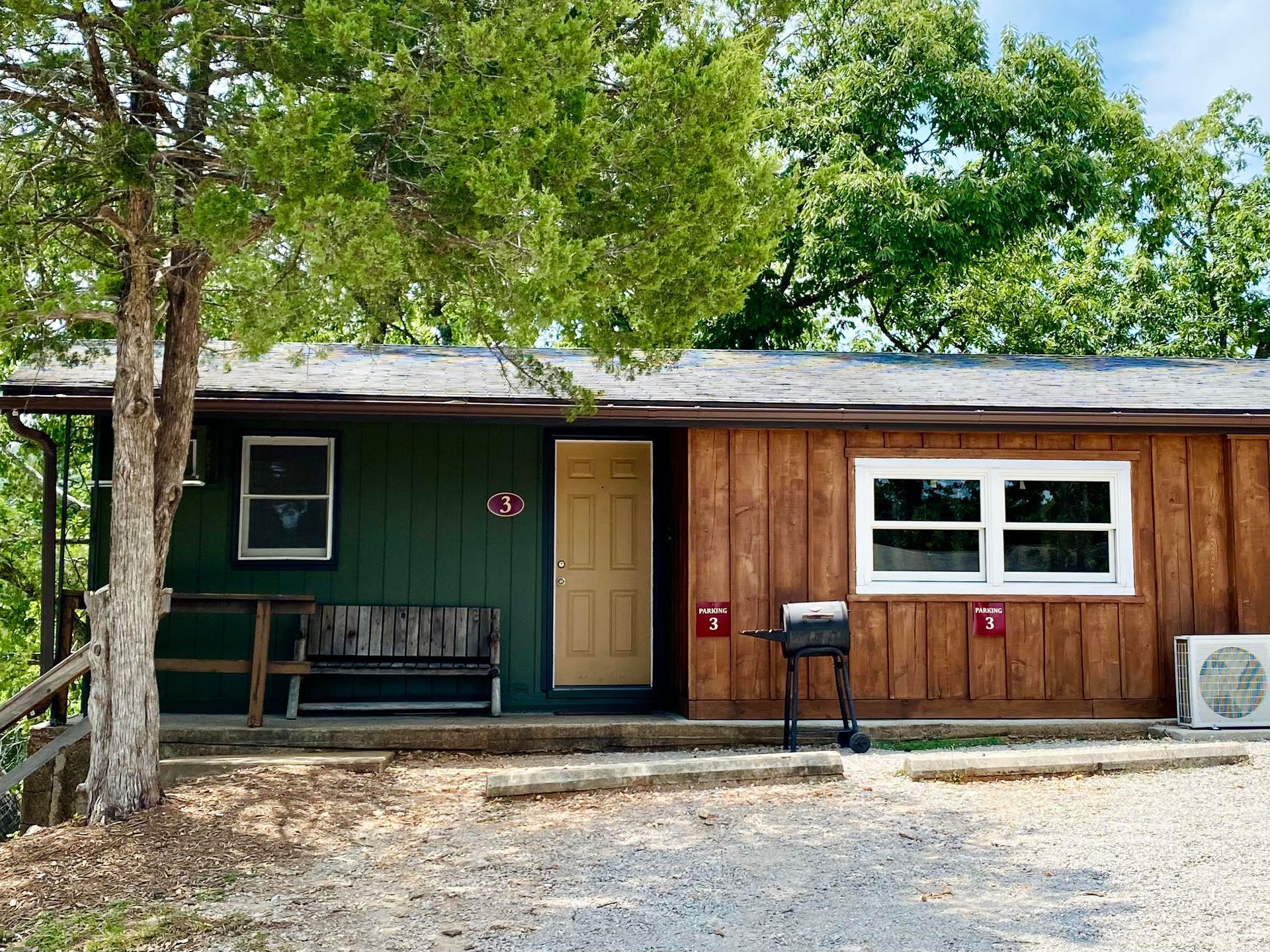 Cabin with green and brown siding, a bench, and a grill. Overcast sky.