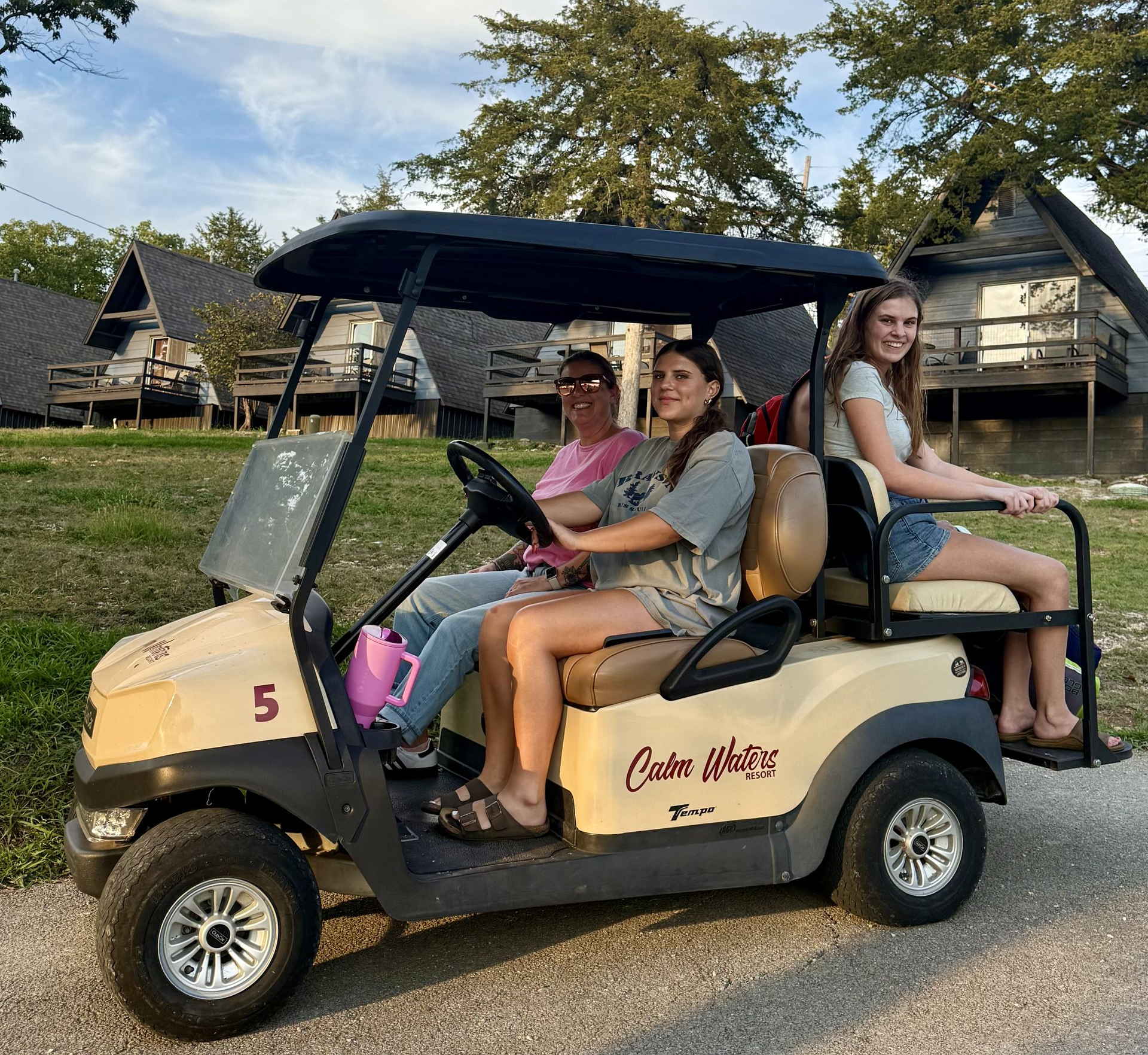 Three people in a beige golf cart on a path, with cabins in the background under a blue sky.