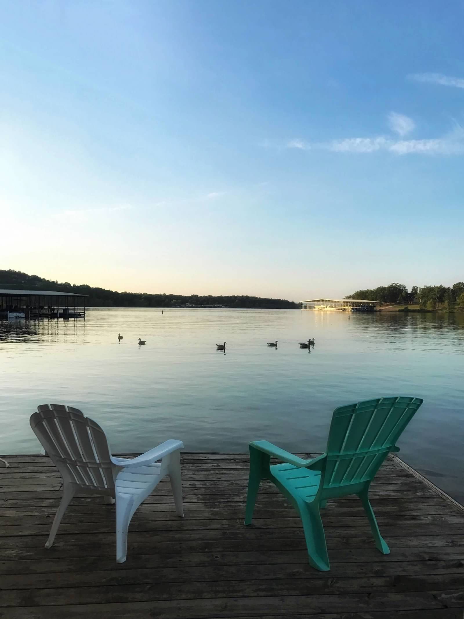 Two chairs on a dock overlook a calm lake with ducks. Blue sky and trees in the distance.