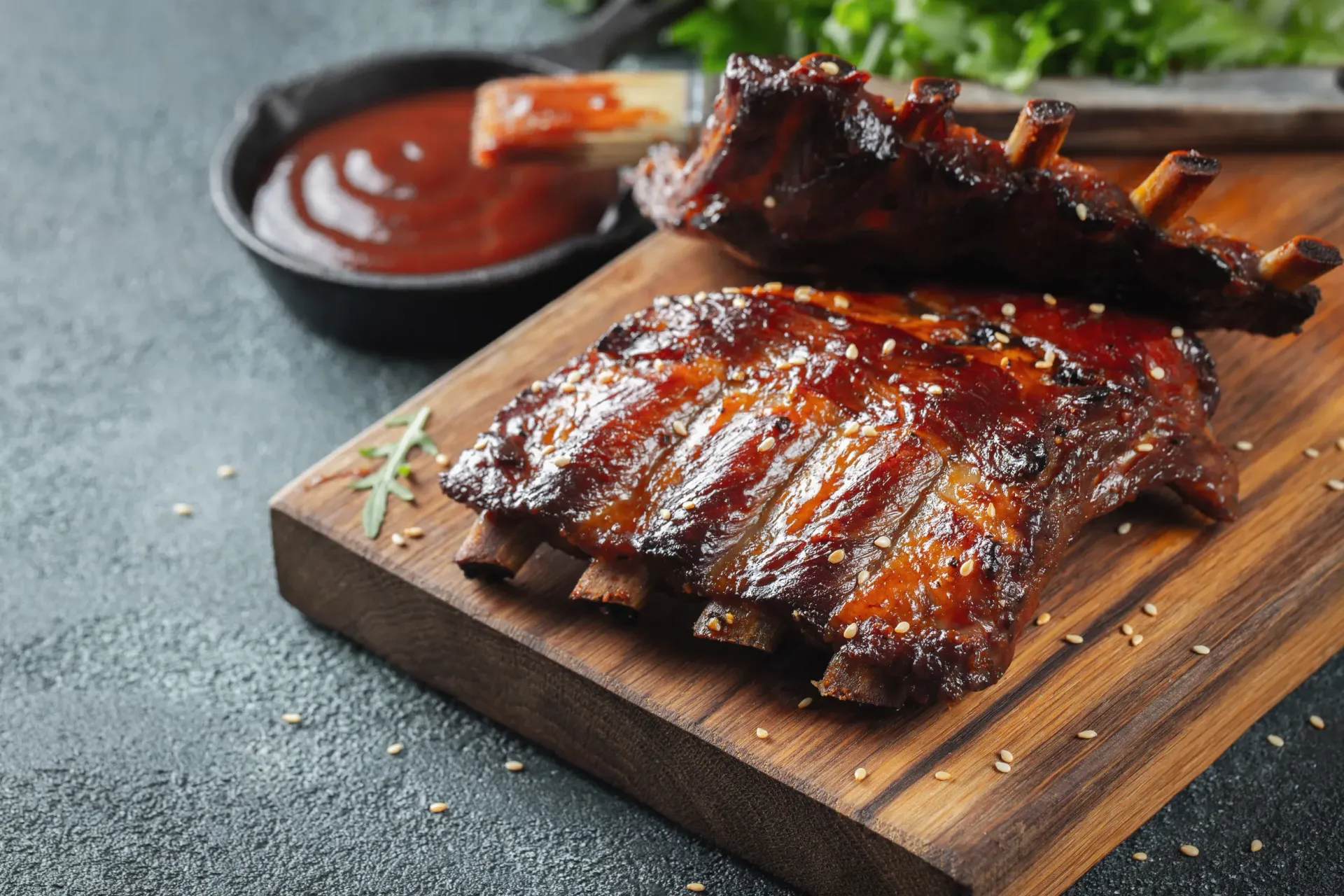 Cooked BBQ ribs on a wooden board, with sauce in a black bowl and greens in the background.