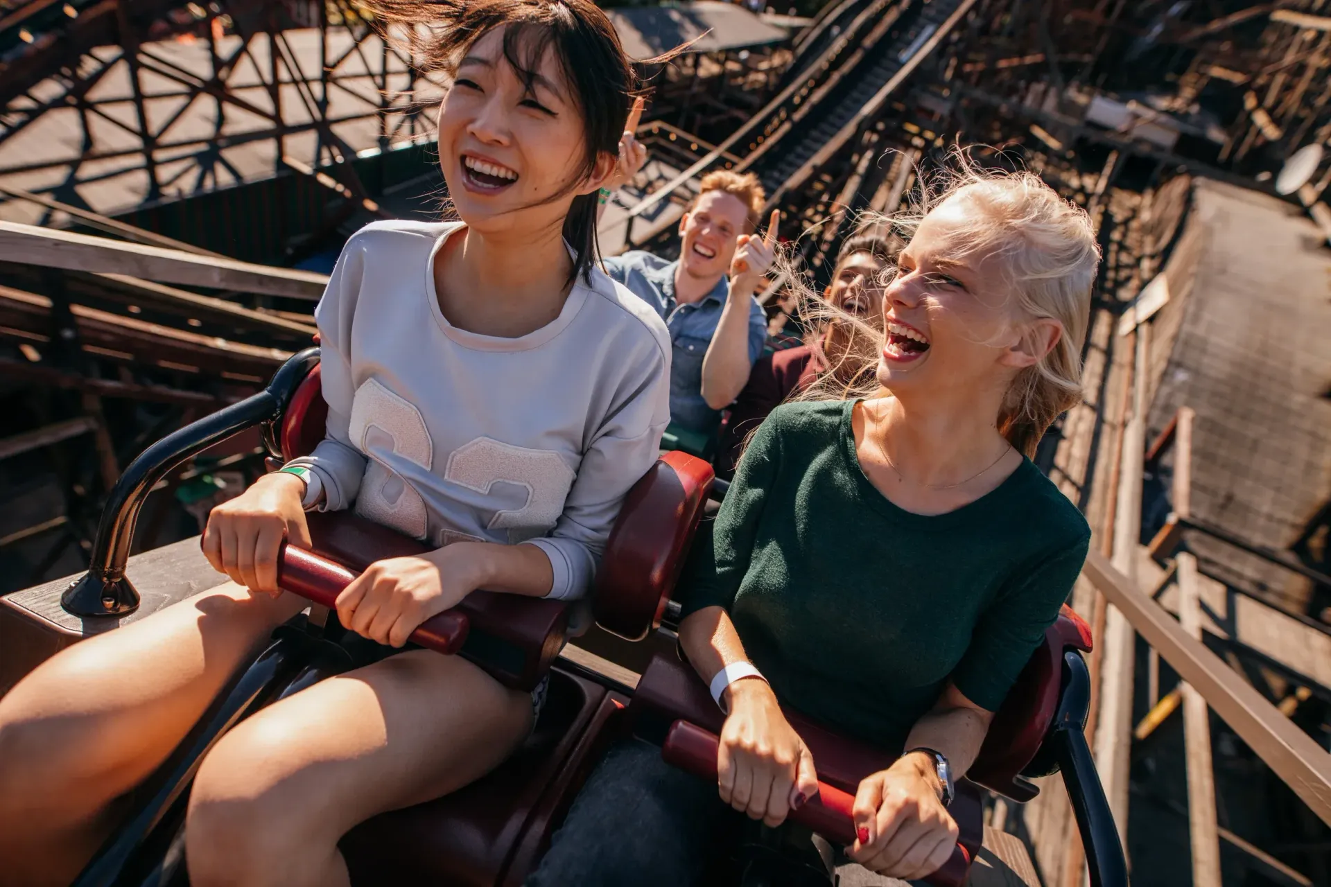 People on a roller coaster, smiling and laughing. Wooden structure visible.