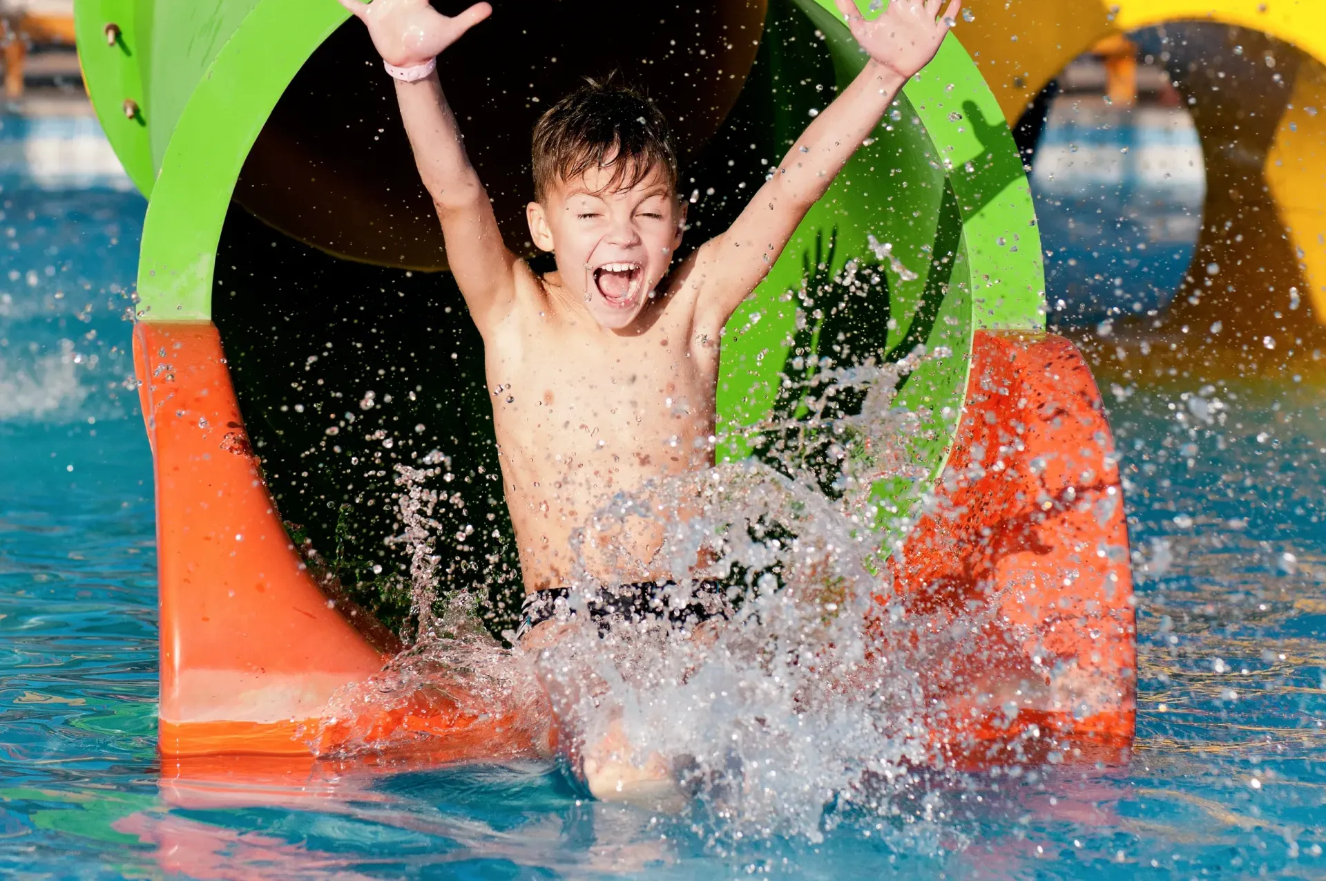 Boy exiting a water slide, splashing into pool. Arms raised, excited expression.