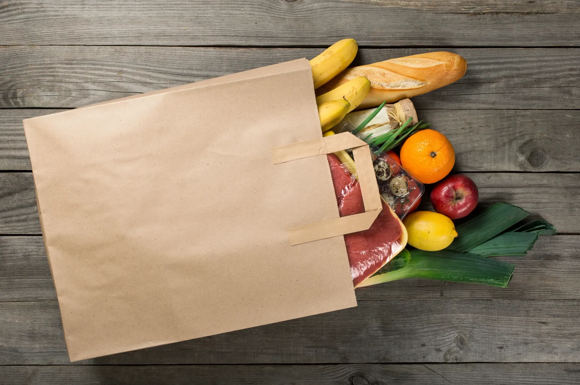 Brown paper grocery bag spilling onto a wood surface, containing fruit, vegetables, and meat.