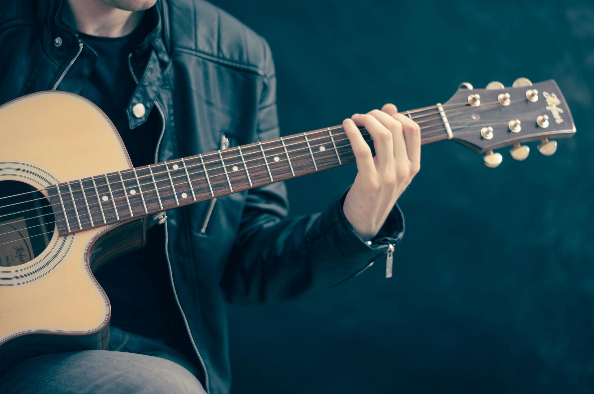 Man playing acoustic guitar, wearing a black jacket, dark background.