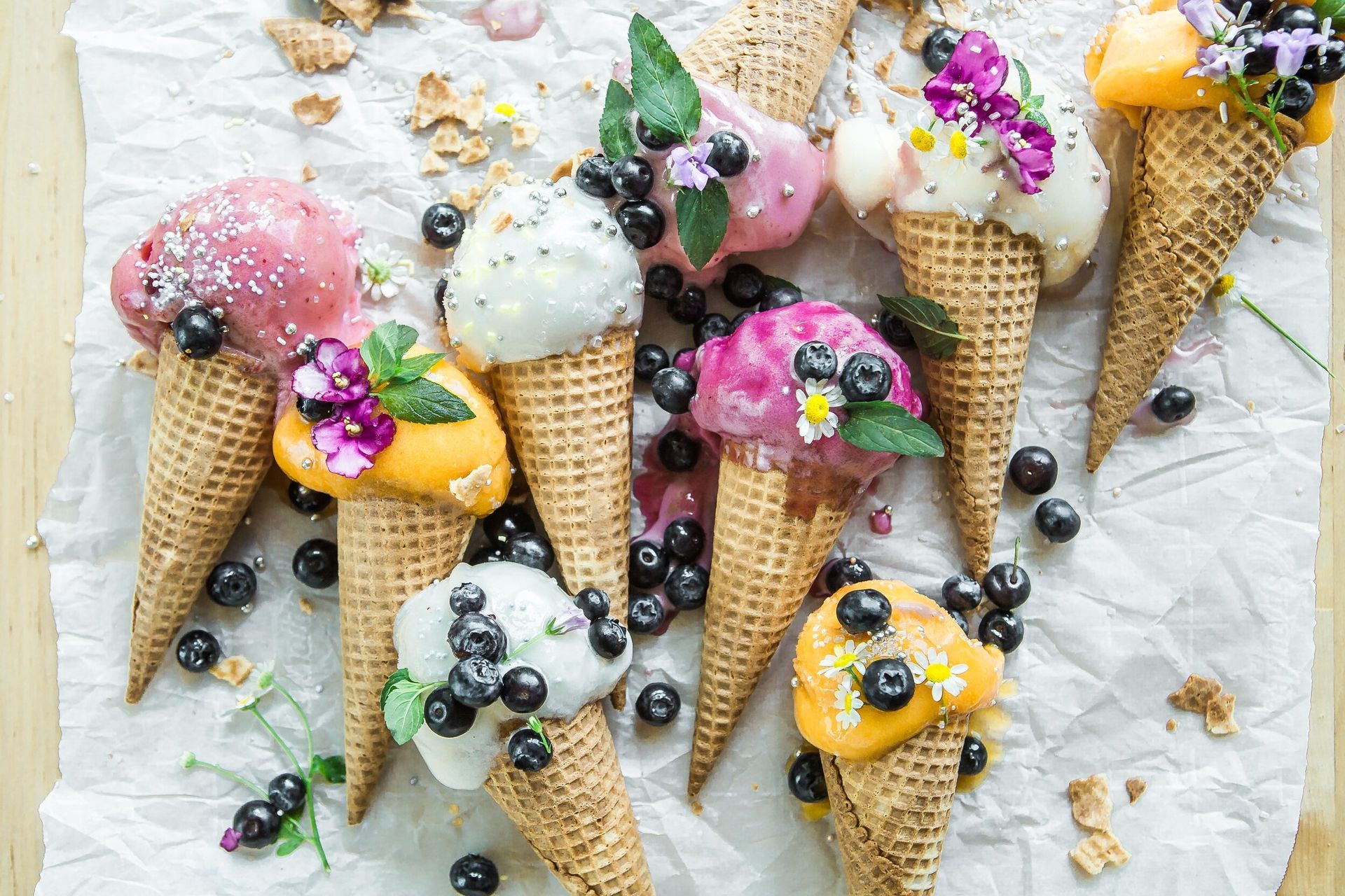 Ice cream cones with colorful scoops, blueberries, and edible flowers on parchment.