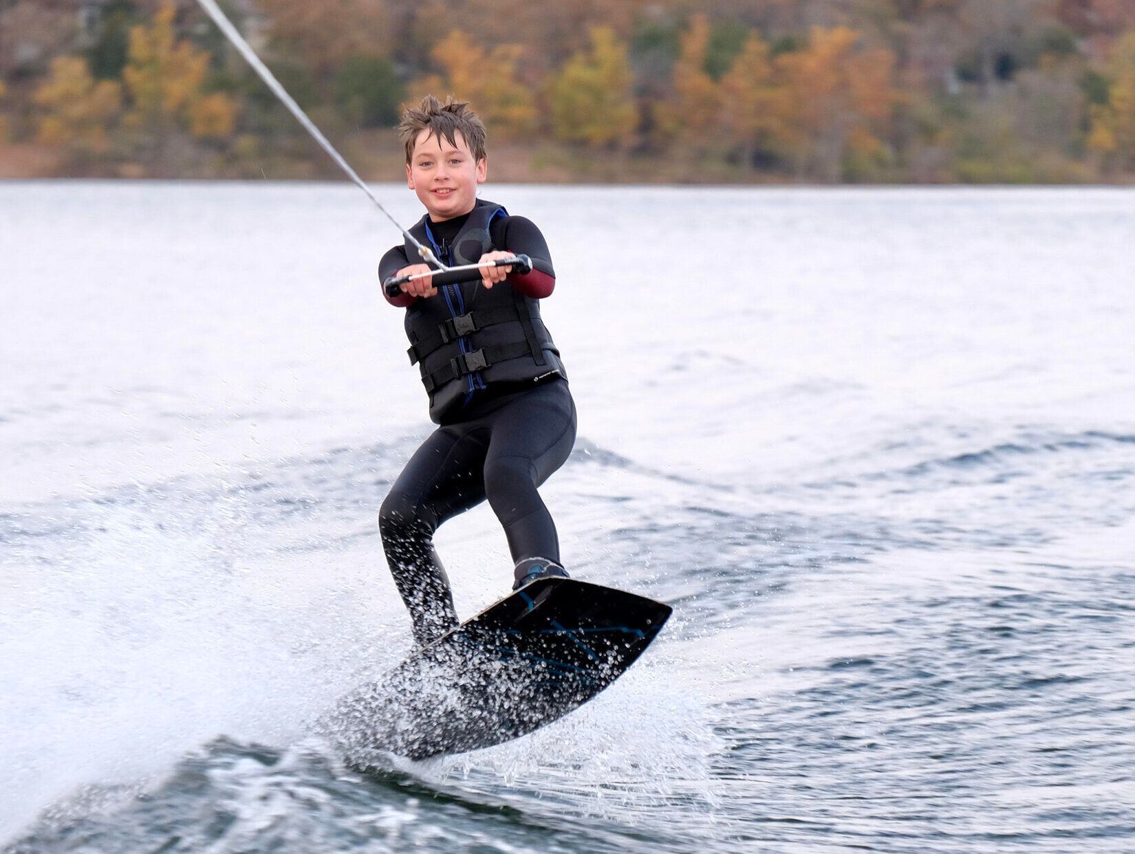 Boy wakeboarding on a lake, holding a rope and handle, wearing a wetsuit and life jacket.