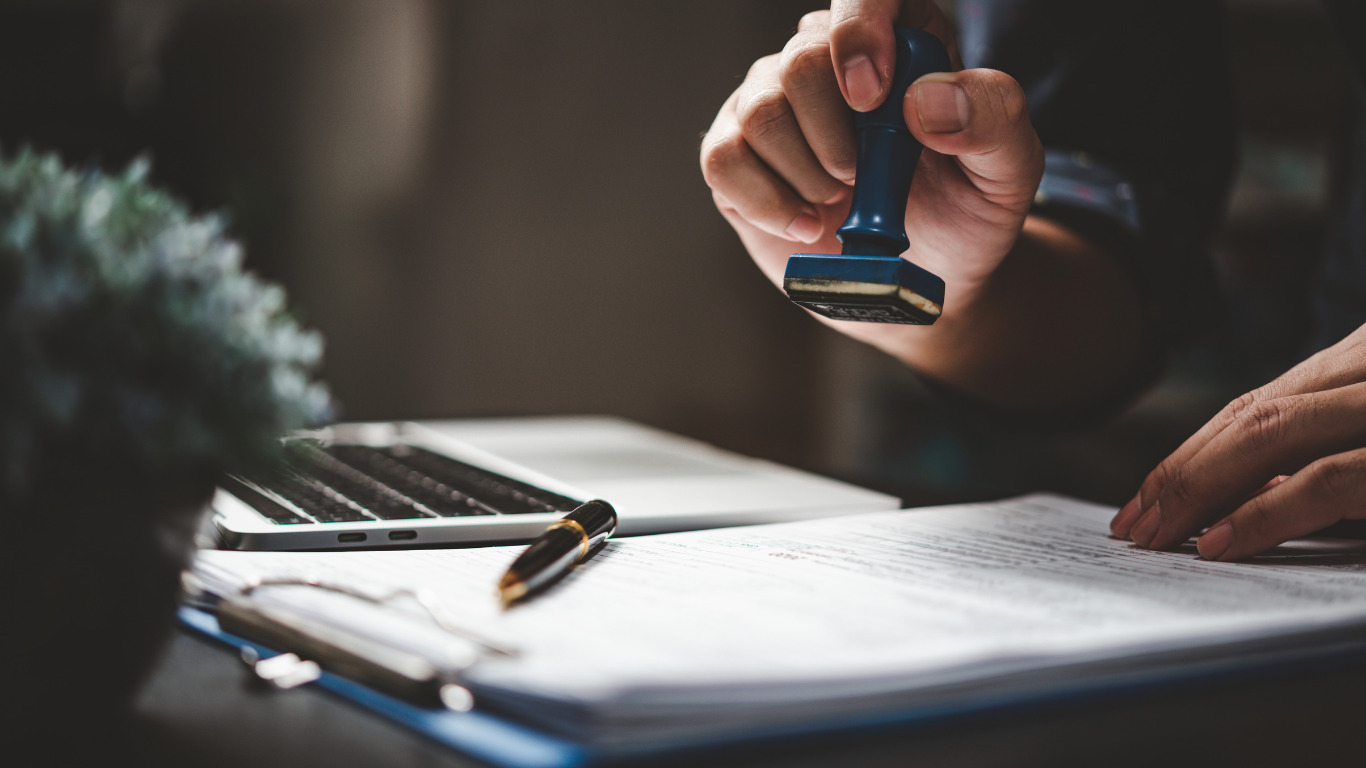 Person stamps a document with a blue stamp near a laptop and pen.