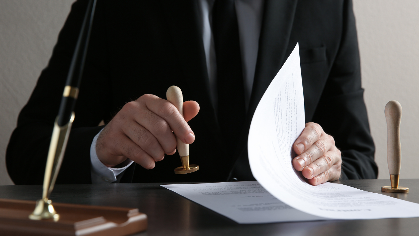 Person in a suit stamping a document with a wooden seal on a desk. A pen is in the foreground.