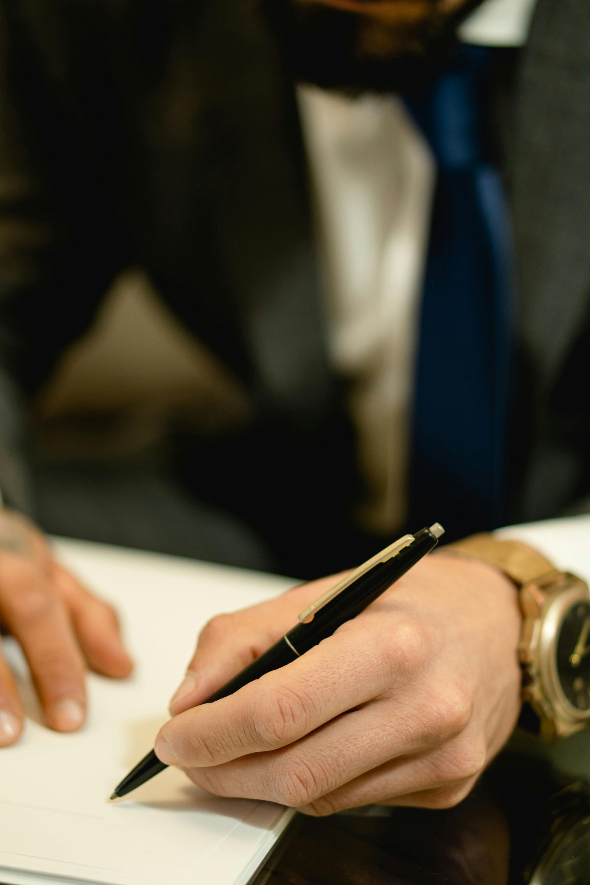 Person in suit writes with a pen on paper, wrist watch visible, close-up.