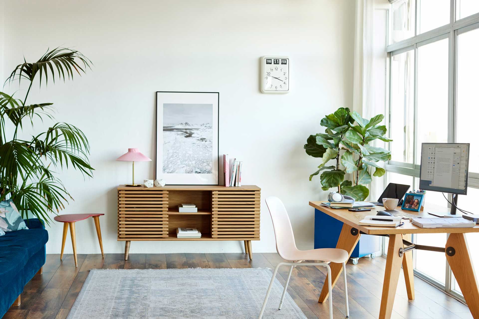 A living room with a desk , chairs , plants and a clock on the wall.
