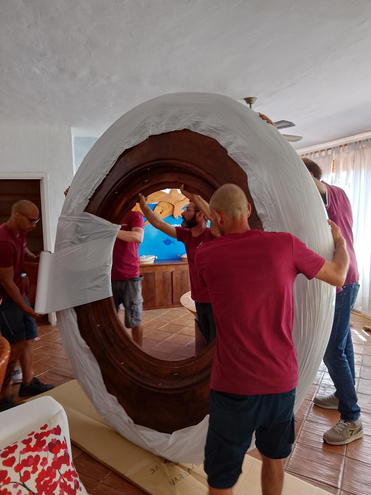 Quattro uomini in camicia rossa spostano con cautela un grande specchio di legno decorato e avvolto in un involucro.