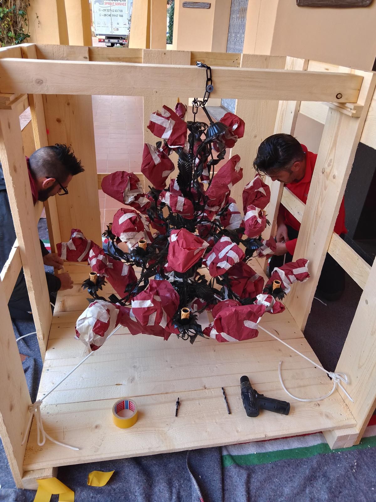 Due persone lavorano su un albero di Natale all'interno di una cassa di legno; decorazioni rosse e bianche.